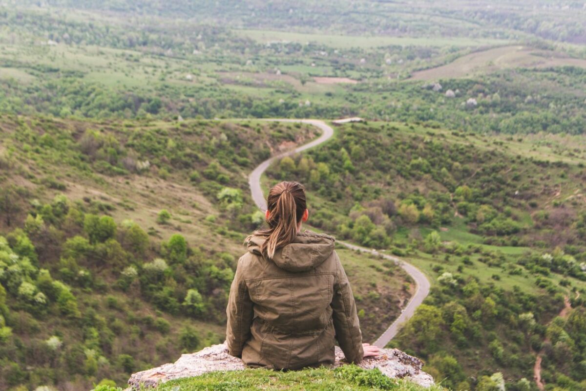 Een vrouw in een bruine hoodie, gezien op de rug, zit op een rots en kijkt uit over een heuvelachtig landschap met een slingerend pad